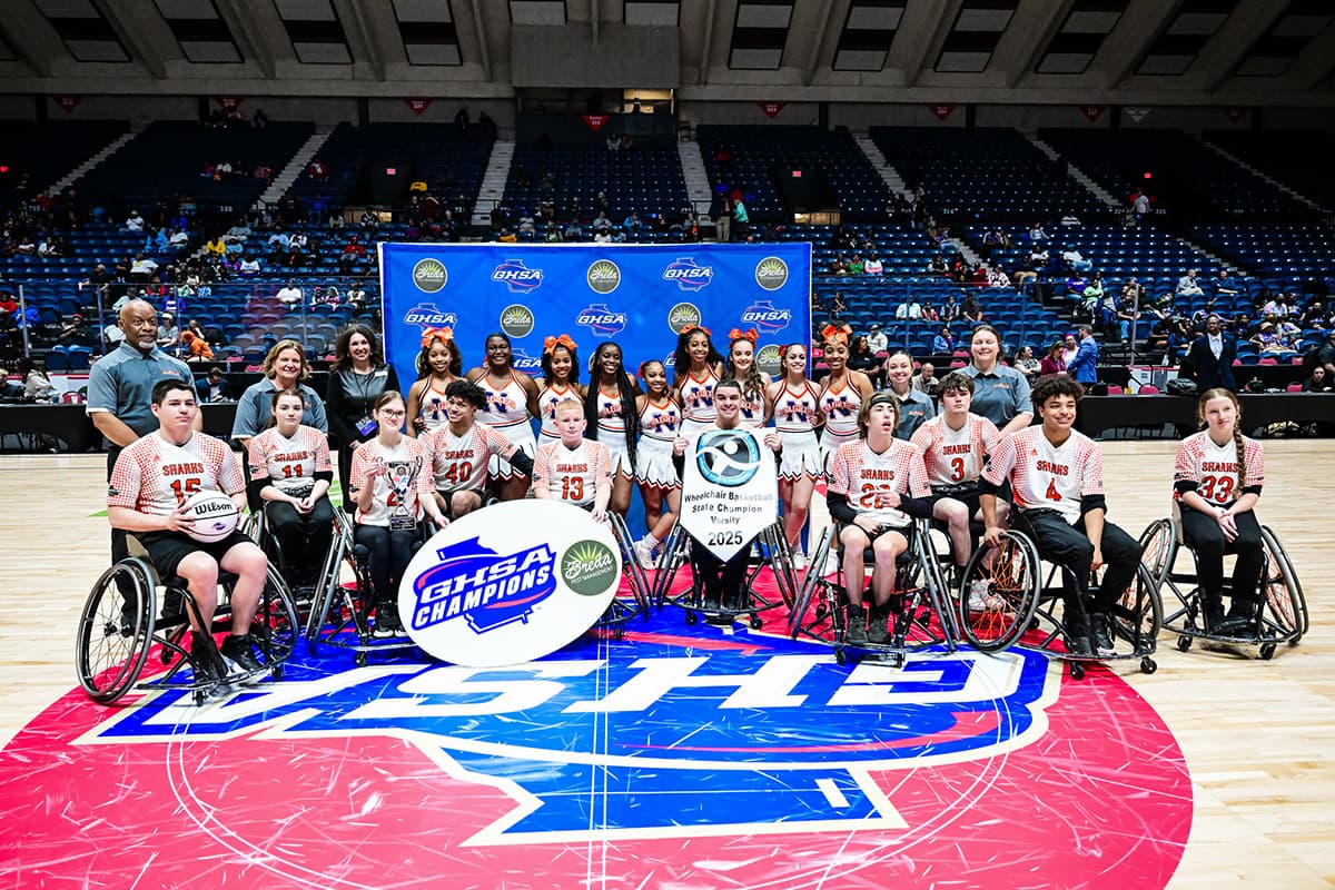 Wheelchair basketball team poses with cheerleaders and a trophy on a court with GHSA banners and logos in the background.