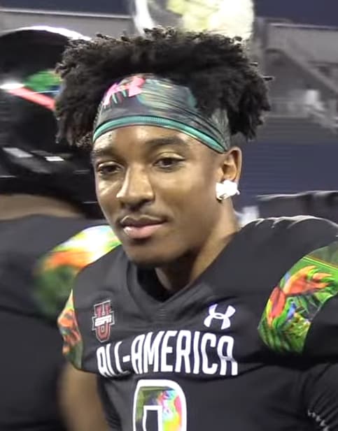 Football player wearing a black "All-America" jersey and colorful headband, standing on the field with a focused expression.